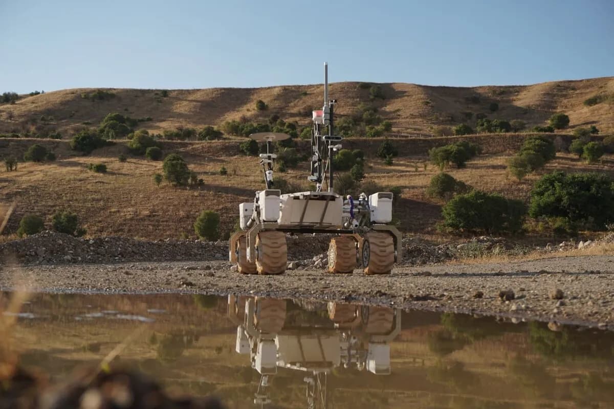 A white rover with large wheels and an antenna stands on rocky ground, reflected in a muddy puddle, with a dry, hilly landscape and blue sky in the background.