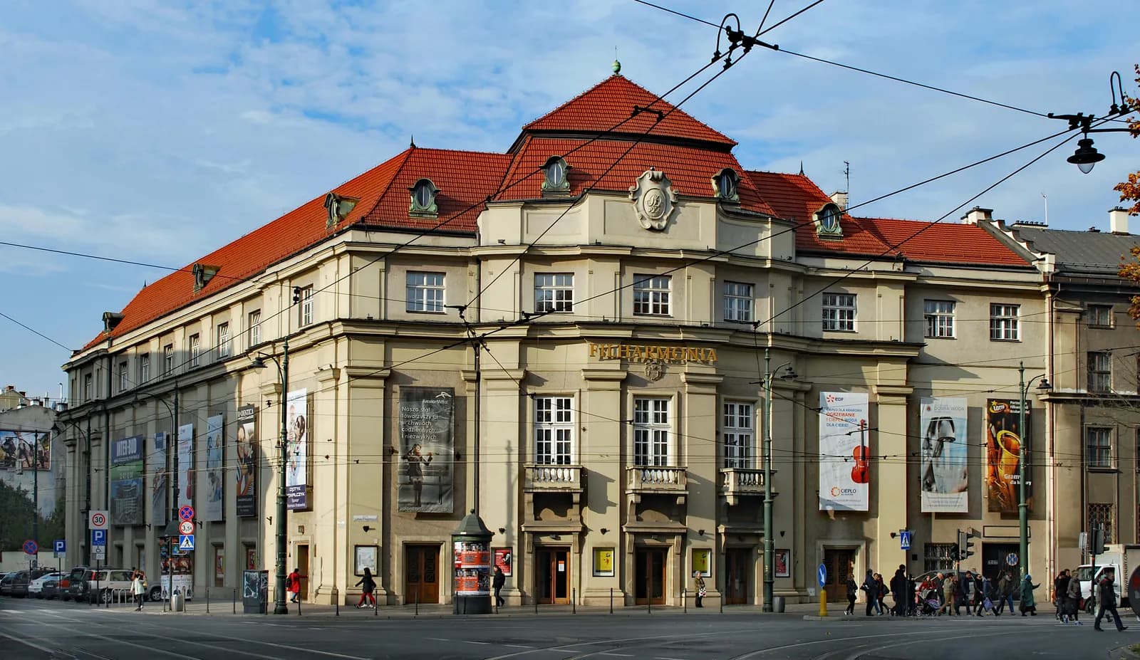A large, multi-story beige building with a distinctive red-tiled roof and numerous windows, featuring banners and advertisements on its facade, stands at a city intersection under a partly cloudy sky.