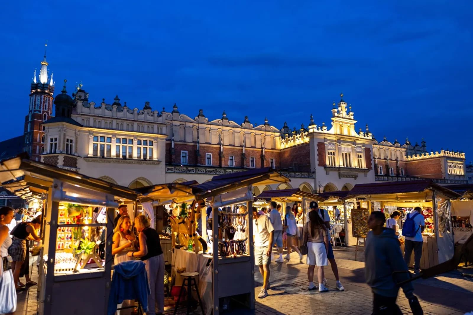 A lively night market with numerous stalls selling goods, bustling with people, set against the backdrop of a grand, illuminated historical building with a prominent tower under a dark blue sky.