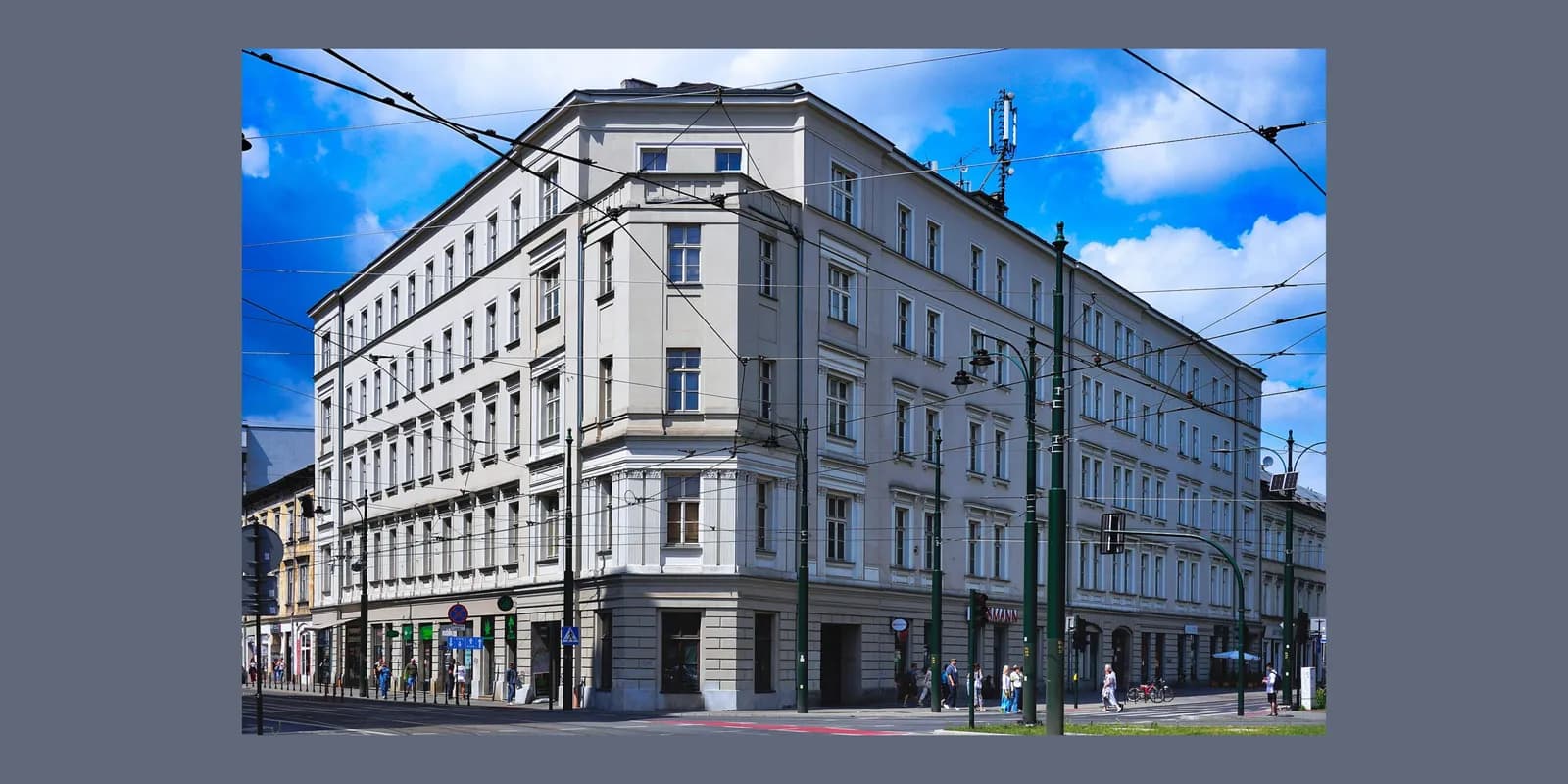 A large, multi-story building with many windows sits at a street corner under a blue sky with white clouds, with overhead wires crisscrossing the frame and people walking on the sidewalks.