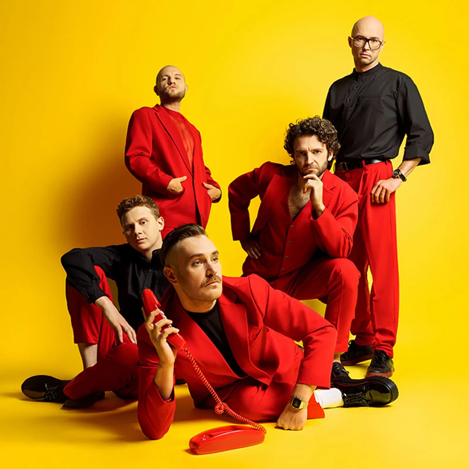 A band of five men in red and black suits posing against a bright yellow background, one man holding a red telephone.